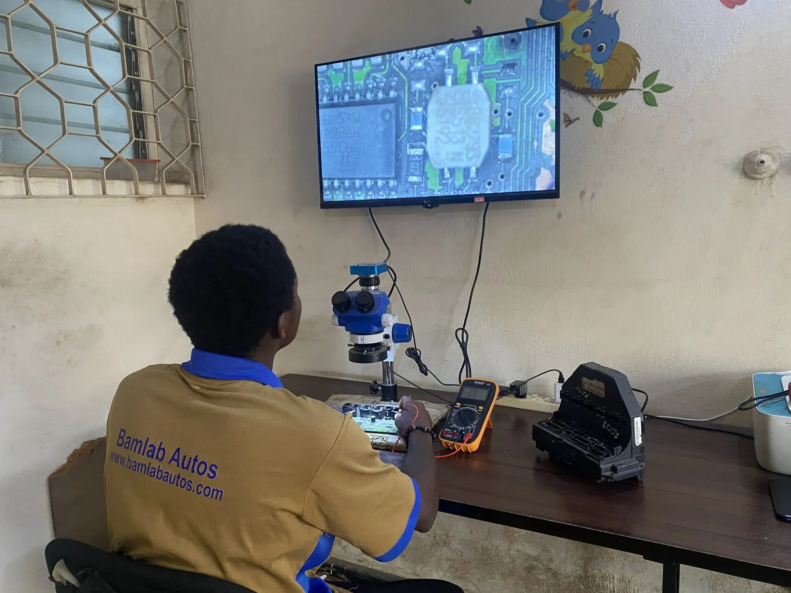 A Bamlab Autos technician repairing a module under a microscope, with the circuit board magnified on a wall screen